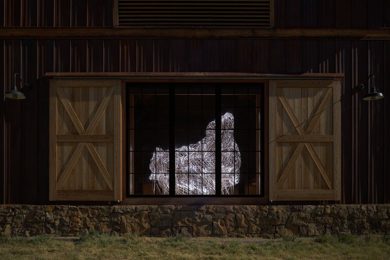 A woven flat reed installation titled Hive: Tippet Rise by Stephen Talasnik inside the Oliver Music Barn gallery space at Tippet Rise Art Center in Montana, viewed through the gallery window at night.